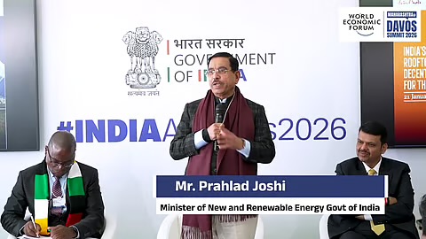 Pralhad Joshi standing and speaking with a microphone at a Government of India pavilion session in Davos, with an on-screen caption naming him as Minister of New and Renewable Energy