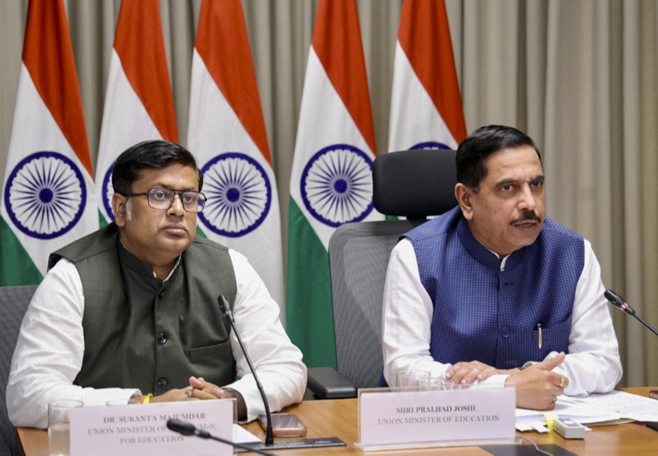 Pralhad Joshi seated at a meeting table with a name plate reading Union Minister of Education, beside Dr Sukanta Majumdar, with Indian flags behind