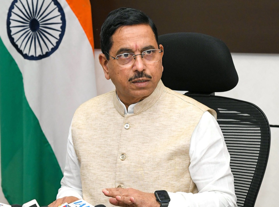 Pralhad Joshi speaking, seated in front of the Indian flag, with media microphones in the foreground