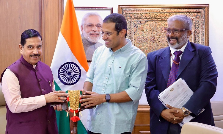 Pralhad Joshi receiving a traditional garland memento from a visitor, with another visitor holding documents beside them