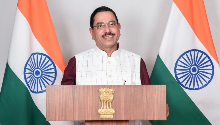 Pralhad Joshi smiling at a wooden lectern with the State Emblem of India, flanked by two Indian flags